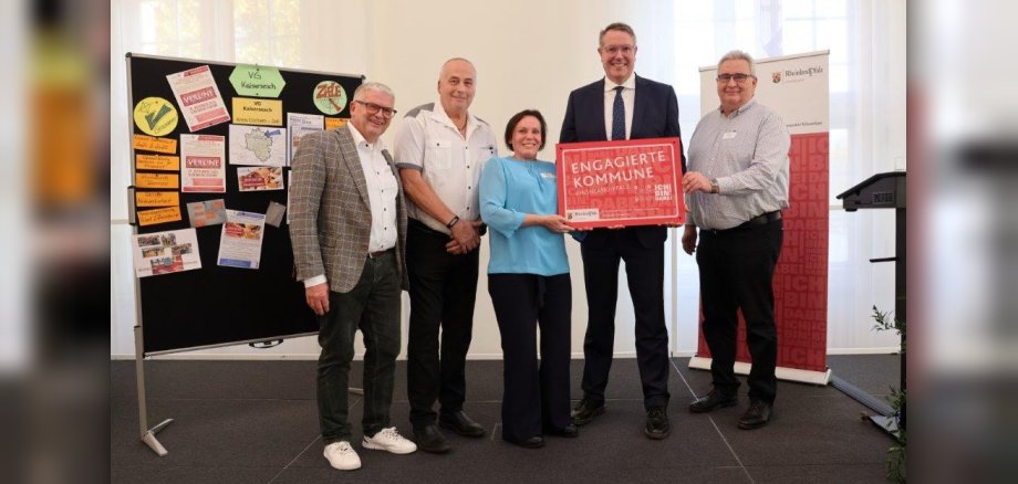 Vor einer Stellwand, die mit vielen unterschiedlichen Plakaten bestückt ist und einem Roll-up der Staatskanzlei Rheinland-Pfalz, stehen von links nach rechts: Erster Beigeordneter Gerhard Weber, Hans Günter Labonte, Bianca Sesterhenn, Ministerpräsident Alexander Schweitzer, Norbert Fuhrmann. Bianca Sesterhenn und Ministerpräsident Alexander Schweitzer halten gemeinsam ein rotes Schild mit der Aufschrift "Engagierte Kommune" in ihren Händen.