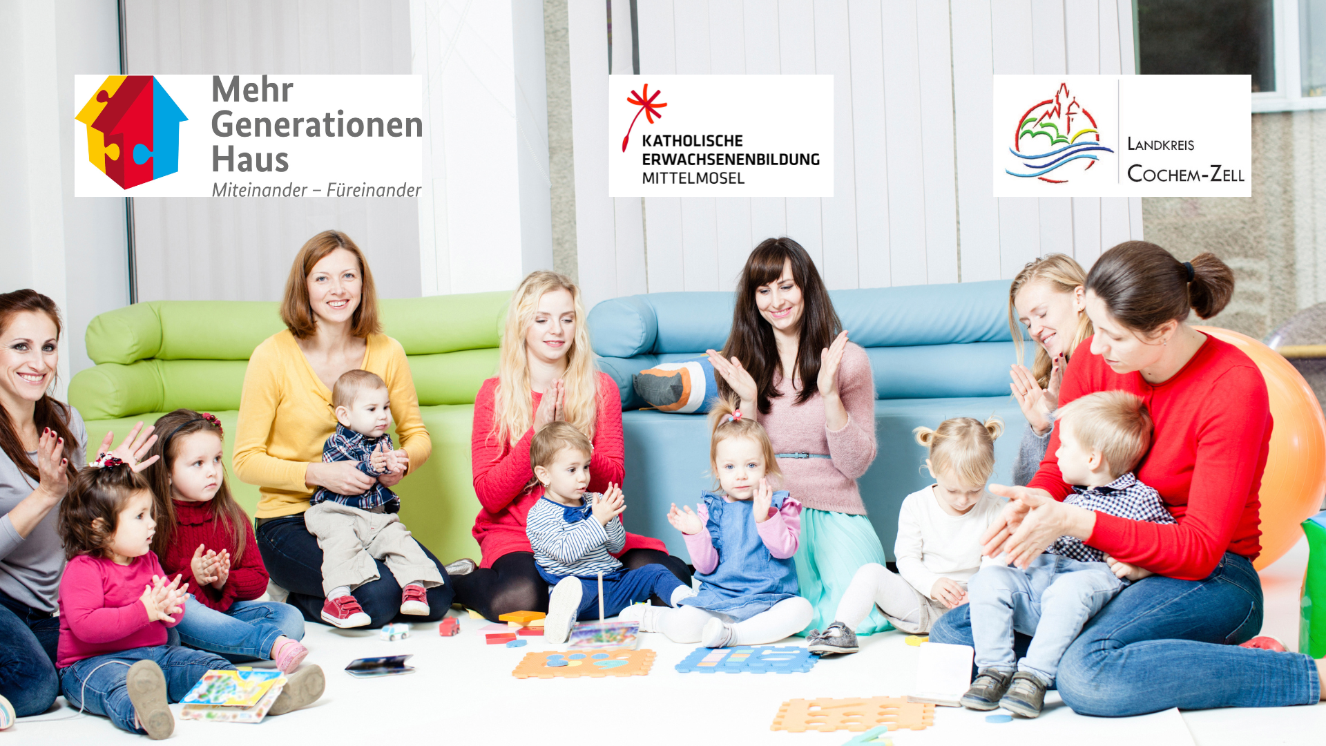Das Bild zeigt eine Gruppe von Frauen und kleinen Kindern, die lächelnd und klatschend auf dem Boden sitzen. Im Hintergrund sind Sofas zu sehen. Oben im Bild sind drei Logos platziert: Eines mit der Aufschrift „Mehr Generationen Haus Miteinander – Füreinander“, ein weiteres mit „KATHOLISCHE ERWACHSENENBILDUNG MITTELMOSEL“ und ein drittes mit „LANDKREIS COCHEM-ZELL“.